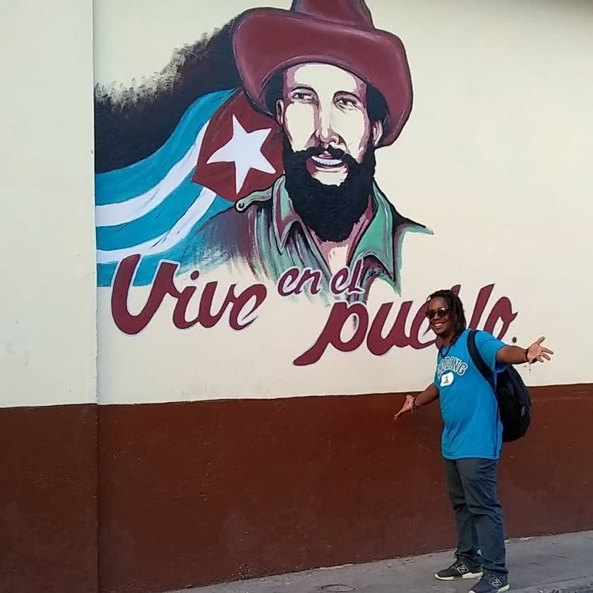Yoyo with open arms in front of the ‘Vive en el pueblo’ mural of Camilo Cienfuegos, 2019